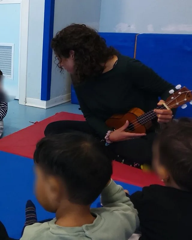 Stefania suona l'ukulele per un gruppo di bambini seduti sui tappeti